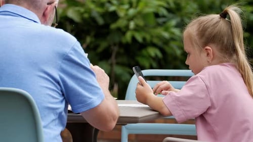 Child Uses Phone at Table Next to Adult