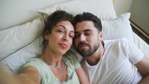 Glückliches amerikanisches Paar macht Selfie und Kuss, während es auf dem Bett im weißen Schlafzimmer liegt Spbd