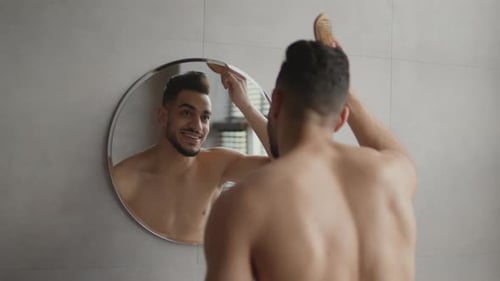 Over Shoulder View of Young Shirtless Middle Eastern Guy Combing His Hair Looking at Mirror at