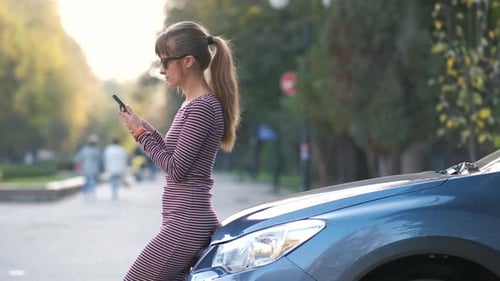 Woman driver standing near her vehicle talking on cellphone on a city street in summer.