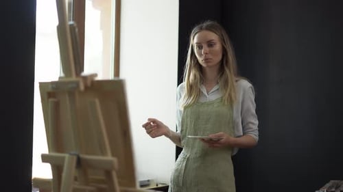 Young Woman Artist in Apron Painting Picture on Canvas in Art Studio