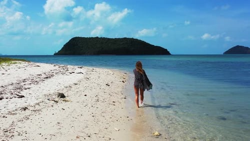 Lonely unrecognizable woman walking on white sandy beach alongside calm clear water of shallow lagoo