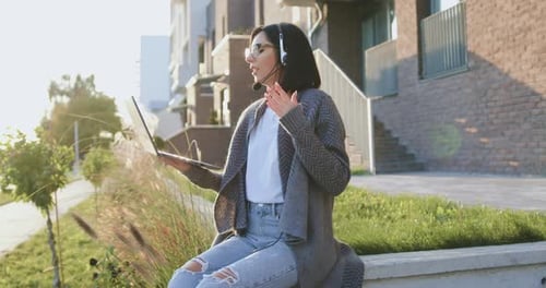 Woman Working with Laptop Device in Headphone Remotely, Sitting on Bench Near Urban street