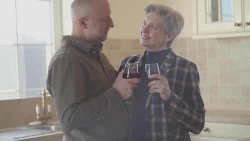 Old Couple Stand in the Kitchen with Wine Glass in Them Hands