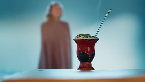 Young Woman Covers Herself with Blanket and Takes Traditional Hot Mate Drink From the Table