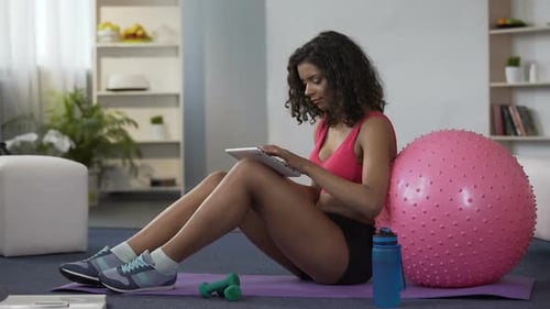 Mixed Race Young Lady in Gym Outfit Sitting on Floor with Tablet, Online Workout