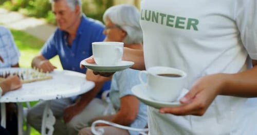 Female volunteer holding cup of tea while seniors playing chess in background 4k