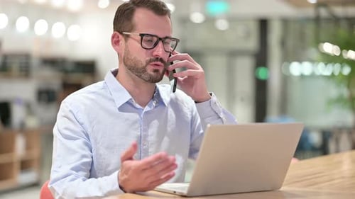 Businessman with Laptop Talking on Smartphone in Office