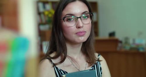 Portrait of Happy Girl in Glasses with Book in Hands Smiles at Camera in Library