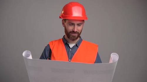 Young handsome civil engineer holding paperwork blueprint and looking at it while standing in studio