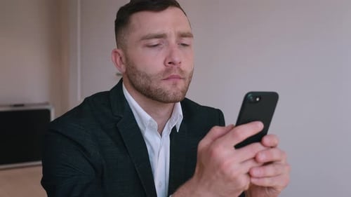 Confident Businessman in Formal Suit Sitting at His Desk in Home Office Using Smartphone, Browsing