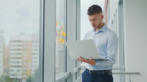 Ein junger Mann hält ein Tablet in den Händen und steht am Fenster. Telearbeit zu Hause und Arbeit in einem