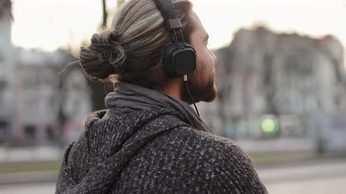 A Bearded Man is Listening to Music with Headphones and Walking Around the City
