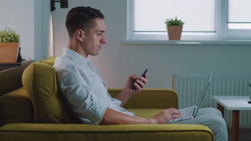 Man Uses Laptop and Phone on Couch Indoors