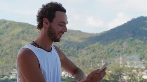 Charmanter junger kaukasischer Mann in einem lässigen weißen T-Shirt steht auf seinem Hotelbalkon