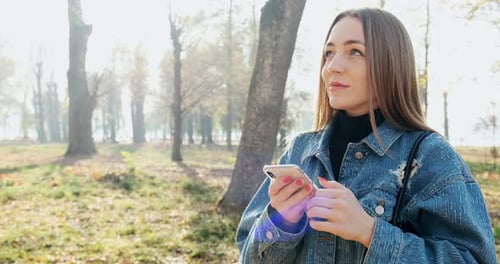 Close Up Portrait of Young Attractive Woman Smiling and Looking at Smartphone at Sunny Autumn Park