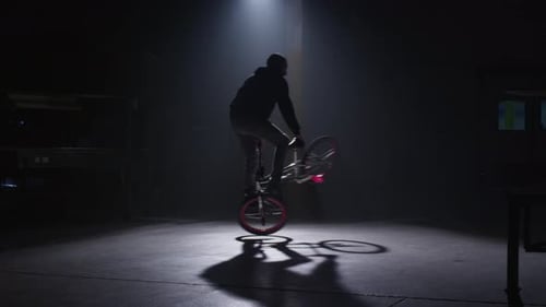 BMX rider doing tricks in dark warehouse.