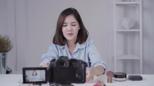 mujer asiática o bloguera de belleza con pincel y cámara grabando Vídeo y agitando la mano en casa.