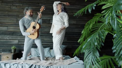 Joyful Couple Dancing on Bed and Laughing While Mature Man Playing Guitar and Singing