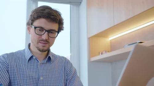 Remotely work on computer. Young man in glasses works at home at a computer, behind him is a window.