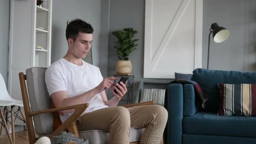 Young Man Using Smartphone Sitting in Chair Indoors