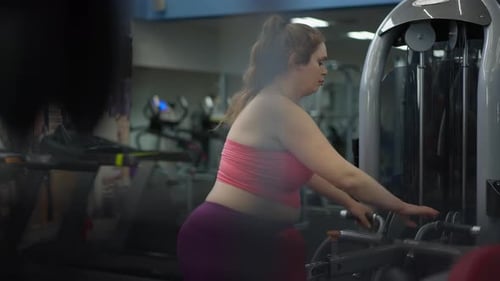 Concentrated Overweight Young Woman Adjusting Exercise Machine with Training Equipment Passing at