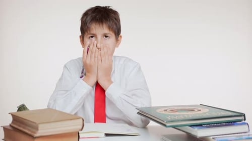 Concentrated Young School Kid in Red Tie Writing Sitting at Table with Books on White Background