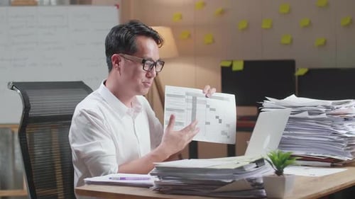Asian Man With Documents Explaining Work On Paper While Having Video Call On Laptop At The Office