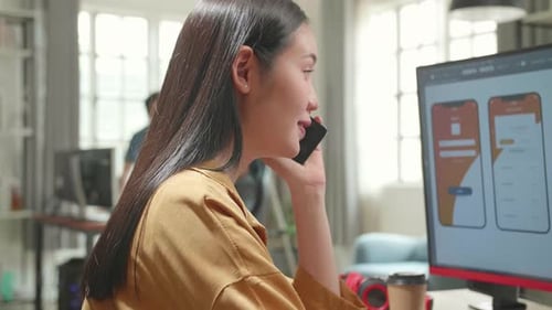 Female Mobile Application Developer Works And Talking On Mobile Phone With Graphics On Computer