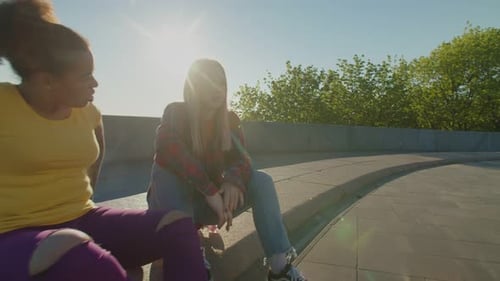 Lovely Chatting Multiracial Women Sitting on Skateboards in City at Sunrise