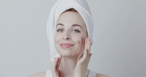 Young Woman Wrapped in White Towel Looking at Camera and Touching Her Clear Perfect Skin