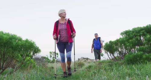 Senior hiker couple with backpacks and hiking poles while walking in the grass field.