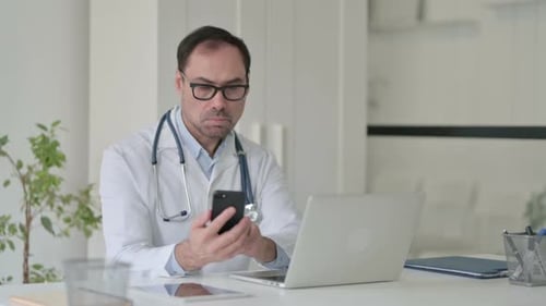 Middle Aged Doctor Using Smartphone While Working on Laptop in Office