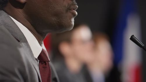 Man Speaking at Press Conference Near Flags