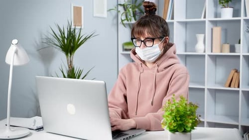 Young woman in protective mask works on laptop at home. Girl student is studying online