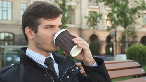 Man Drinks Coffee on Park Bench in City