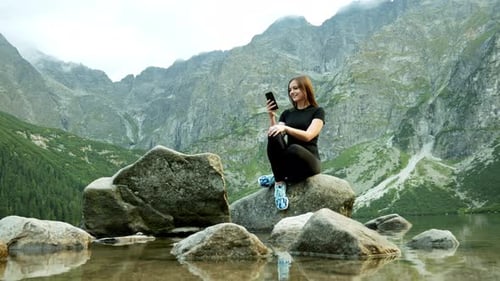 Beautiful Woman Tourist Uses a Mobile Phone While Sitting on a Stone Near a Lake in the Rocky