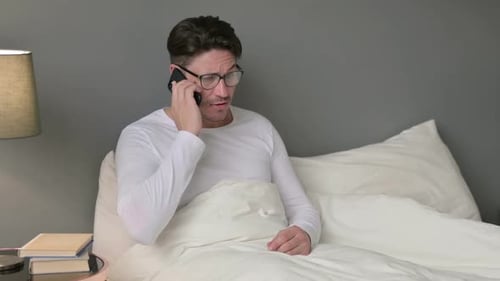 Homme d'âge moyen en colère parlant sur un smartphone au lit