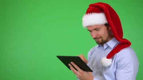 A Young Man in a Christmas Hat Works on a Tablet, Then Smiles at the Camera - Green Screen Studio