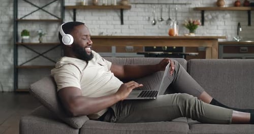 Side View African American Man Entrepreneur Wearing Headphones Talking Video Call at Home Lying on