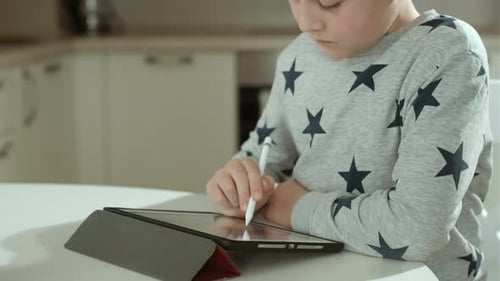 Concentrated boy in grey sweatshirt studying or drawing on tablet at home.