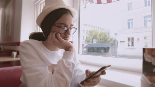 Girl in a Hat and Glasses is Sitting in a Cafe Using Her Cell Phone Waiting for Her Order