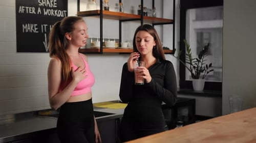 Two female athletes make a protein shake in the gym bar and drink it