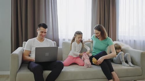 Family Sitting on Couch Together Man Typing on Laptop Woman and Girl Playing Toys Little Boy