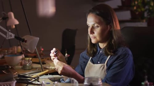 Profile of focused caucasian female jeweller sitting at desk, making jewelry in workshop