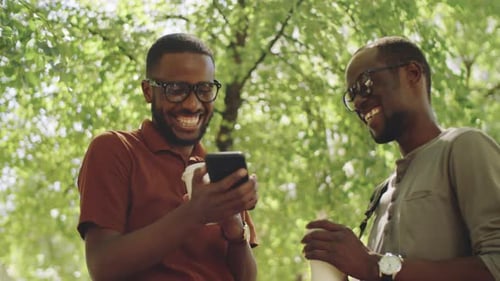 Cheerful Black Male Friends Using Smartphone and Laughing in Park