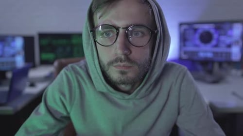 Gros plan d'un homme concentré à lunettes regardant une caméra avec plusieurs ordinateurs au