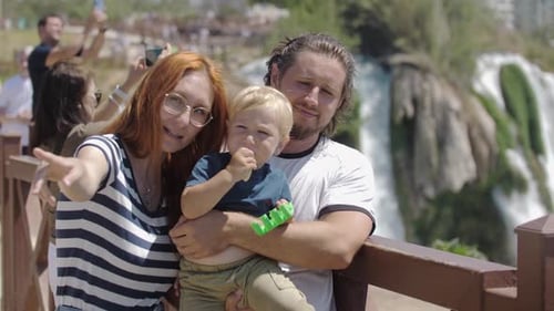 Young Family of Tourists Standing on the Bridge By the Waterfall Father Holding His Son on His Hands