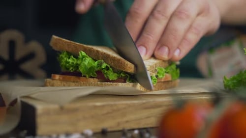 Hombre corta el sándwich del club por la mitad con un cuchillo afilado en una tabla de madera en la cocina