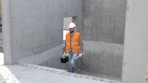 Builder repairman in protective helmet and vest stands at workplace in building and holds ruler